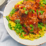 lamb shanks sit on herb almond couscous on a white plate with silverware sitting on a wooden table.