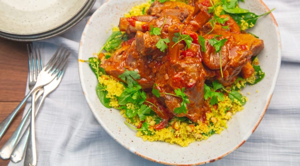 lamb shanks sit on herb almond couscous on a white plate with silverware sitting on a wooden table.