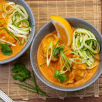 Two bowls of yellow curry topped with green vegetables and a lemon wedge on bamboo mat.