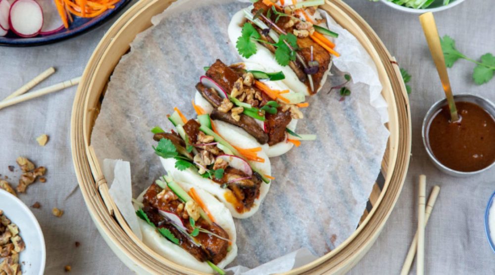 Top view of a bamboo steamer lined white paper and 4 bao buns filled with brown filling on it. Surrounded by chopsticks and small bowls filled with food.
