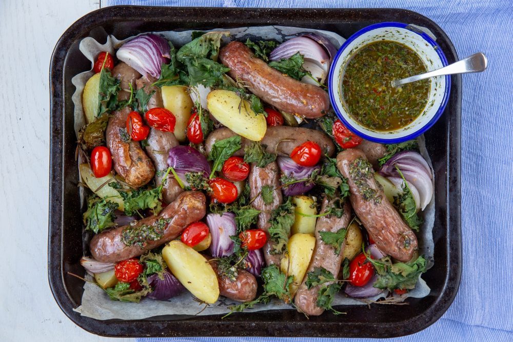 Oven tray with sausage and potato bake. salsa verde in awhite bowl with blue rim and a silver spoon in it. The bake also has onions and herbs sprinkled over.