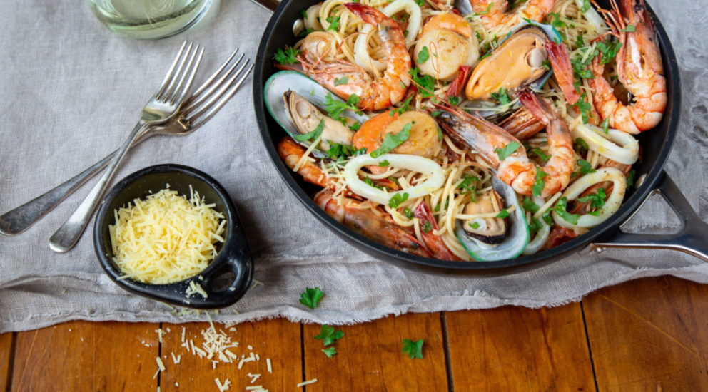 A frypan filled with cooked seafood on white cloth with small dish of grated cheese and forks