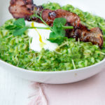 Green rice topped with a cooked bacon and white sauce in a white bowl on pink cloth and a spoon.