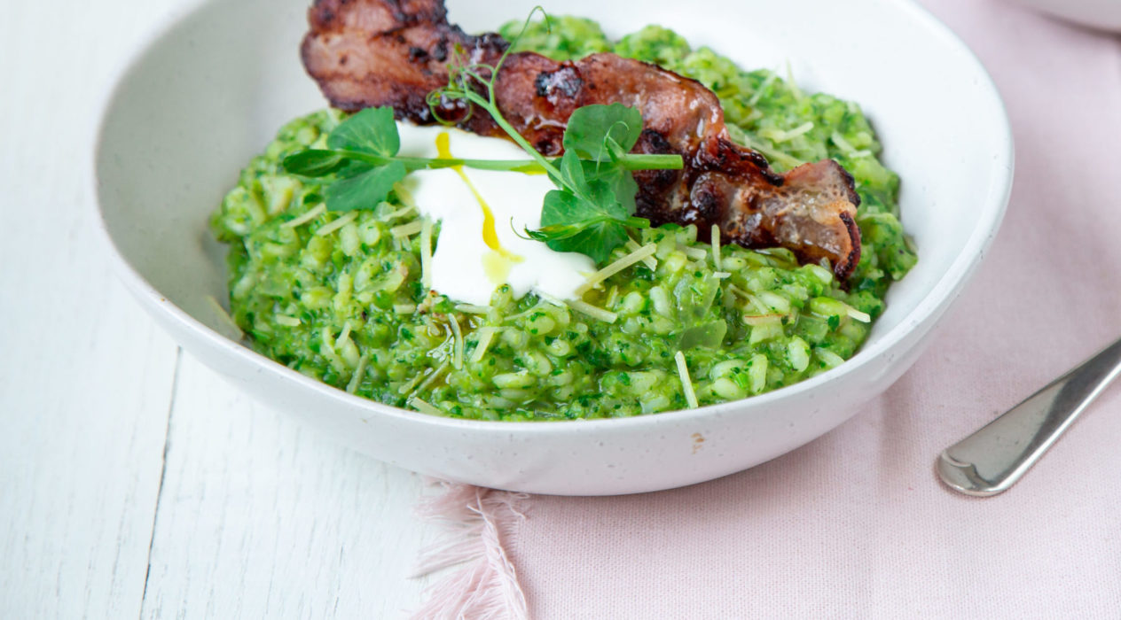 Green rice topped with a cooked bacon and white sauce in a white bowl on pink cloth and a spoon.