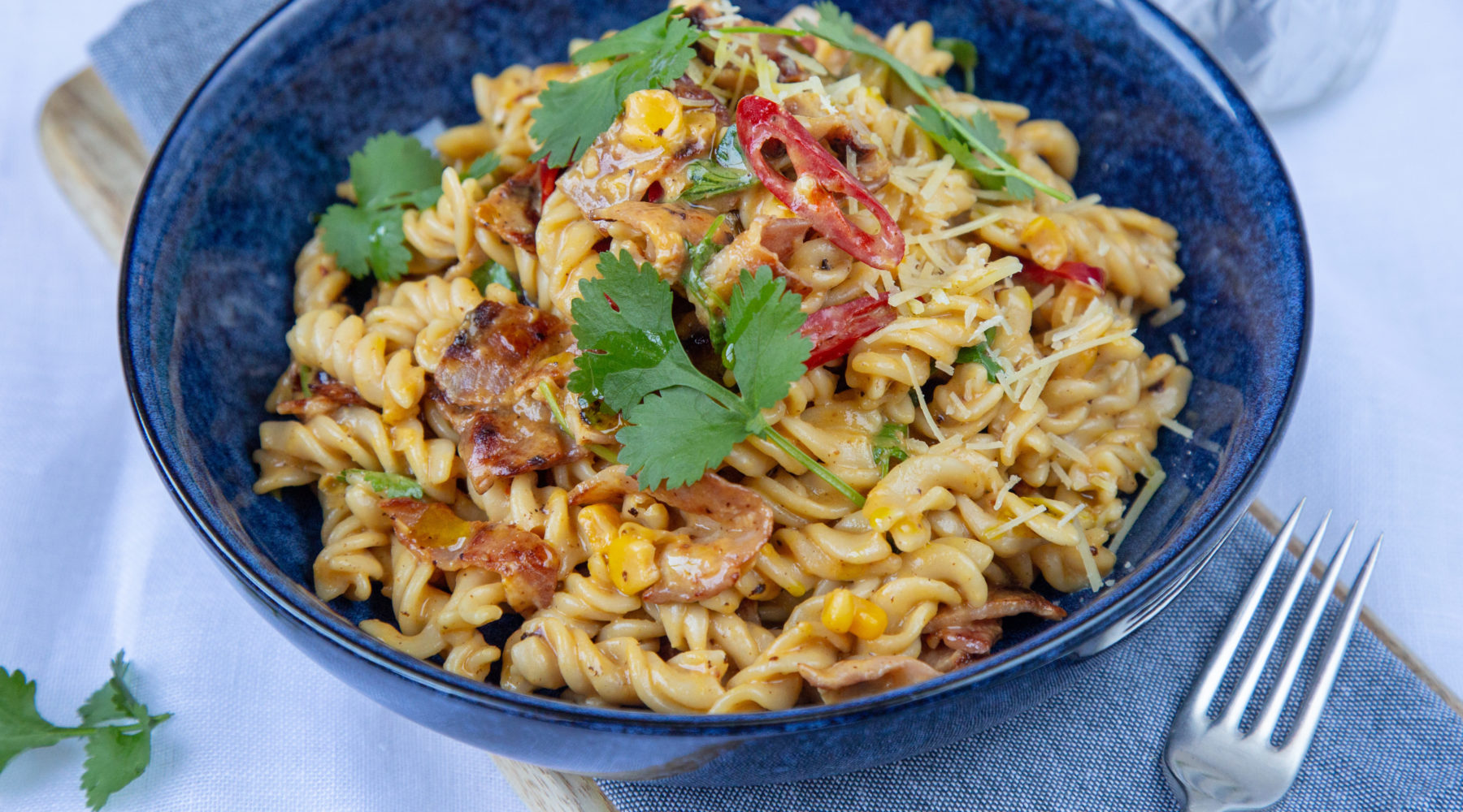 A blue bowl full of pasta with red and green food topped on light blue cloth with a fork.