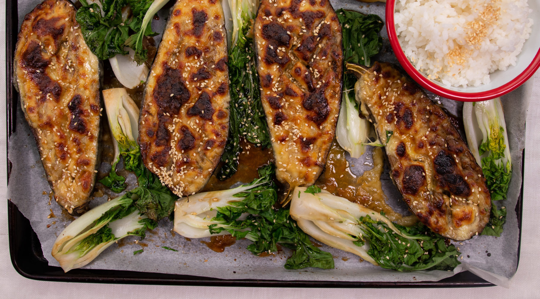 Four halves of aubergine pasted with sauce and greilled with steamed Asian greens and a bowl of rice on metal tray.