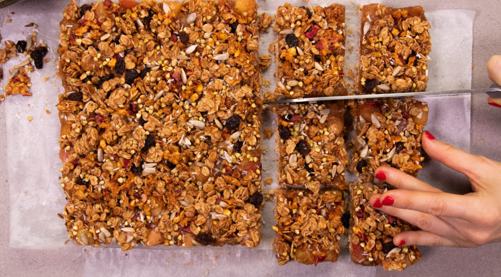 A top view of a large granola slice, a hand holding right bottom side and knife cutting into it just above.
