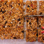 A top view of a large granola slice, a hand holding right bottom side and knife cutting into it just above.