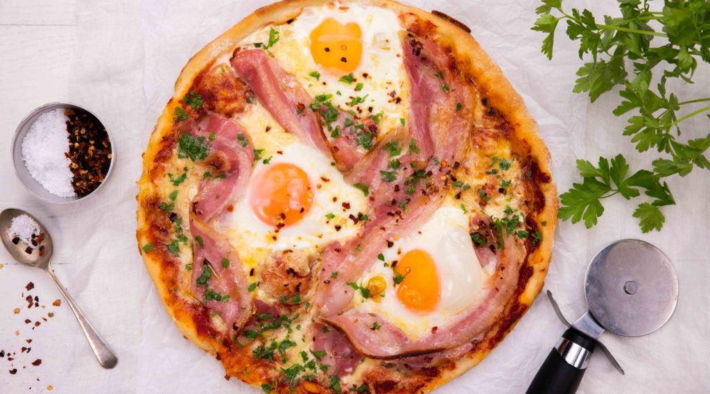3 eggs and bacon strips on a pizza and a pizza cutter, spoon & green herb.