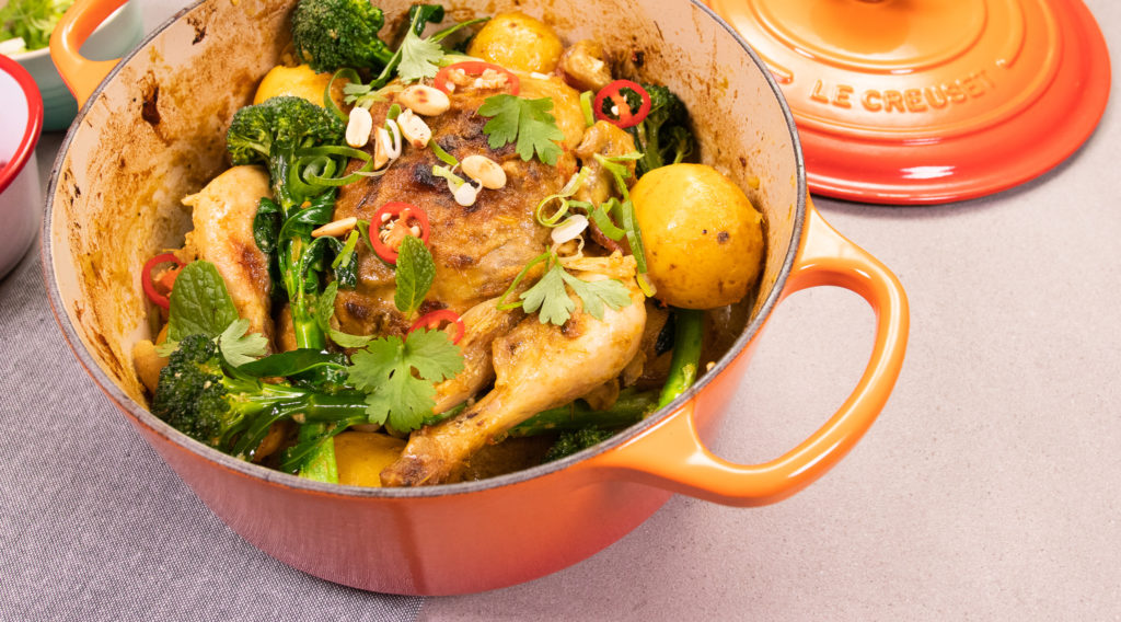 A orange casserole pan with a lid, filled with a whole cooked chicken topped with greens and chill