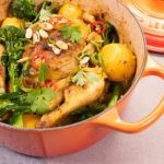 A orange casserole pan with a lid, filled with a whole cooked chicken topped with greens and chill