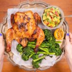 Hands holding a silver tray with whole roast chicken,broccoli, lemon half and a pot of salsa on it over wooden board