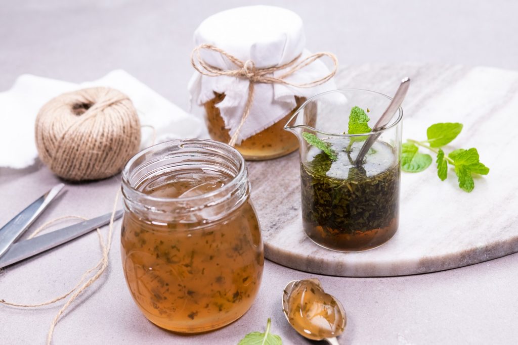 2 jars of brown jelly with one still open top and a cup of dark green liquid with spoon. Fresh mint, a string ball and scissors.