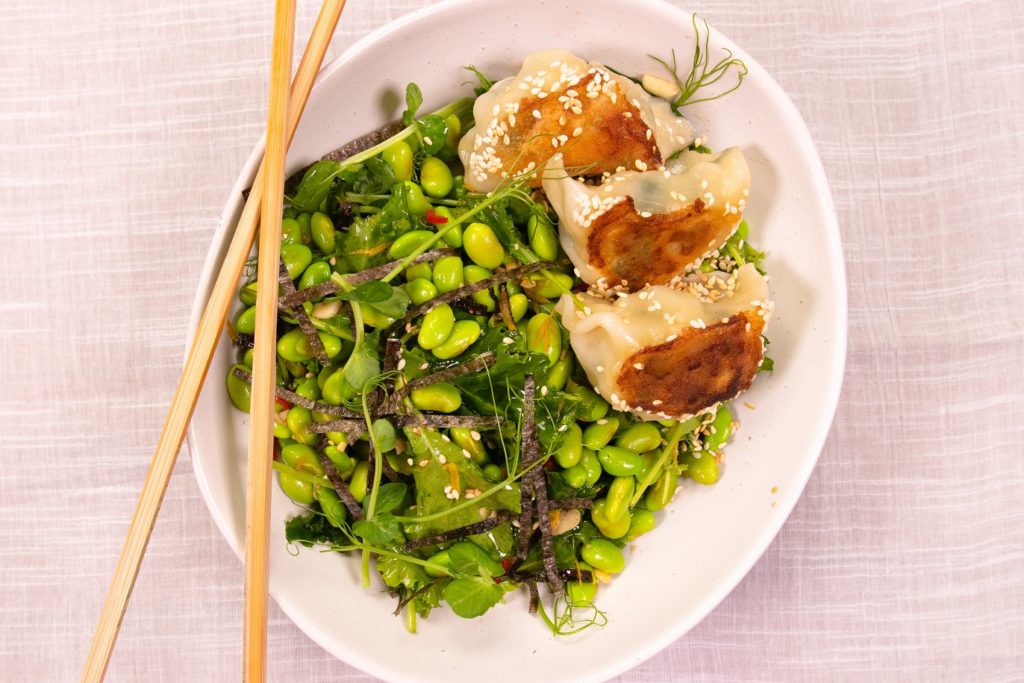 3 dumplings on bed of green bean salad in an oval white bowl with chopsticks on pale pink cloth.