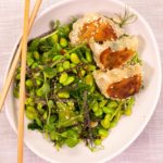 3 dumplings on bed of green bean salad in an oval white bowl with chopsticks on pale pink cloth.