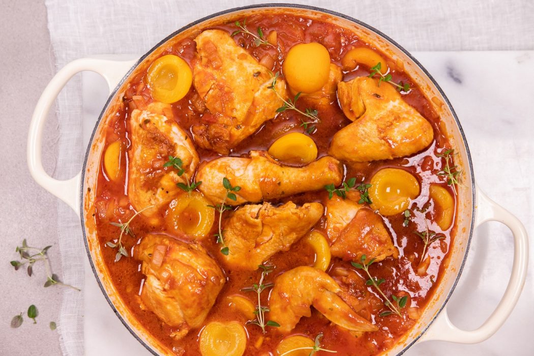 Chicken pieces and apricot halves in reddish brown sauce in a white casserole pan with handles on white board