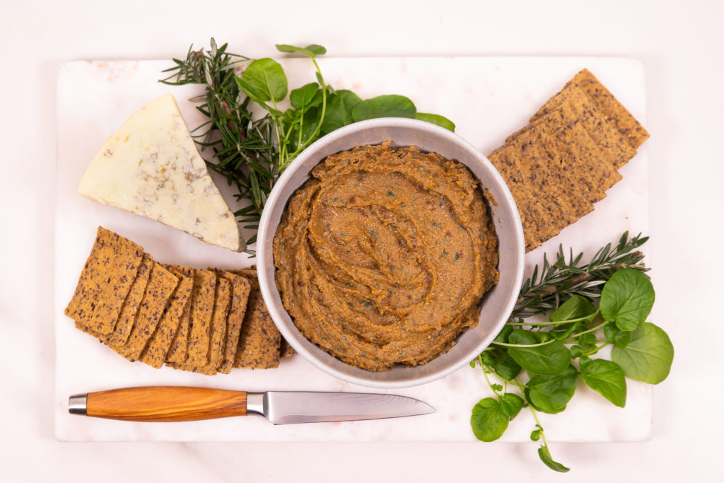 A round bowl of brown pate surrounded by a knife, seed crackers, blue cheese, herbs on a white board.