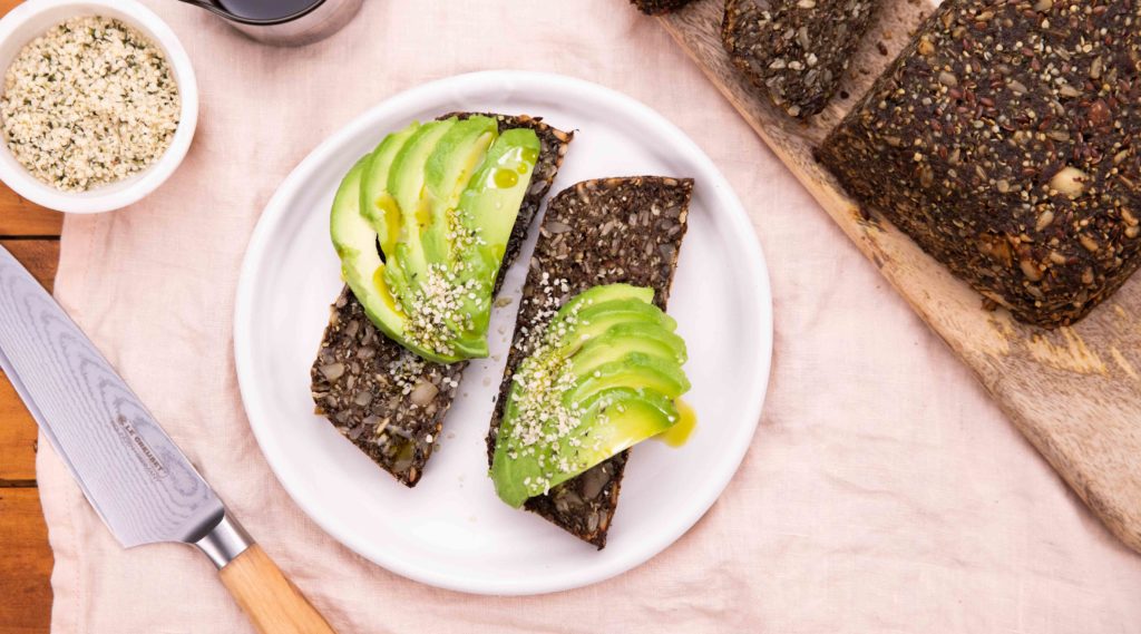 2 slices of dark brown bread with avocado slices on top on a white round plate next to the brown loaf on wooden board with a knife and a small pots of seeds
