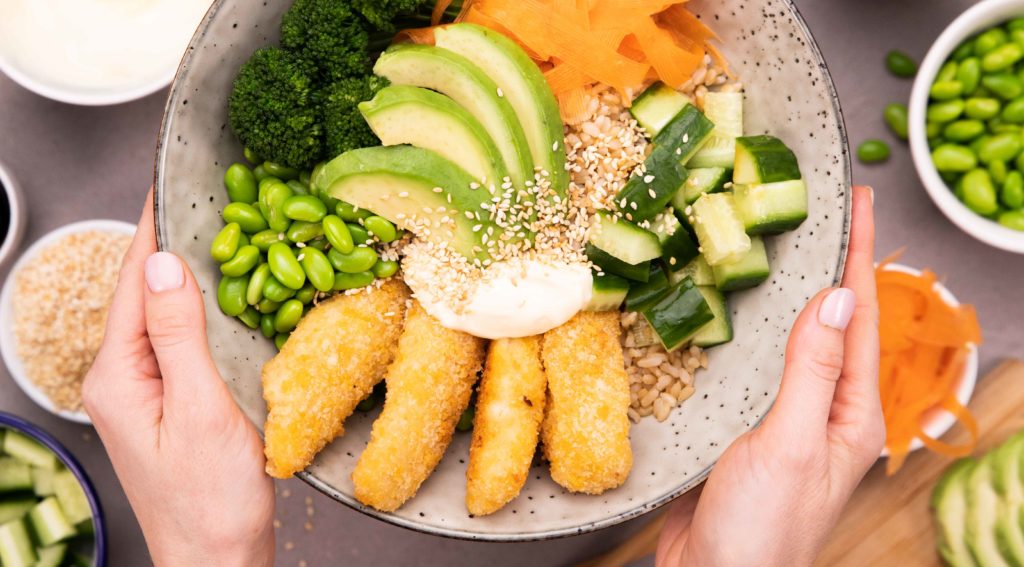 Hands holding a large shallow bowl with fish nuggets and vegetables over small bowls of vegetables