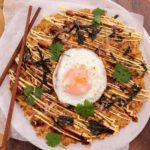 A brown pancake topped with a fried egg and pices of nori & herb and chopsticks on wooden board