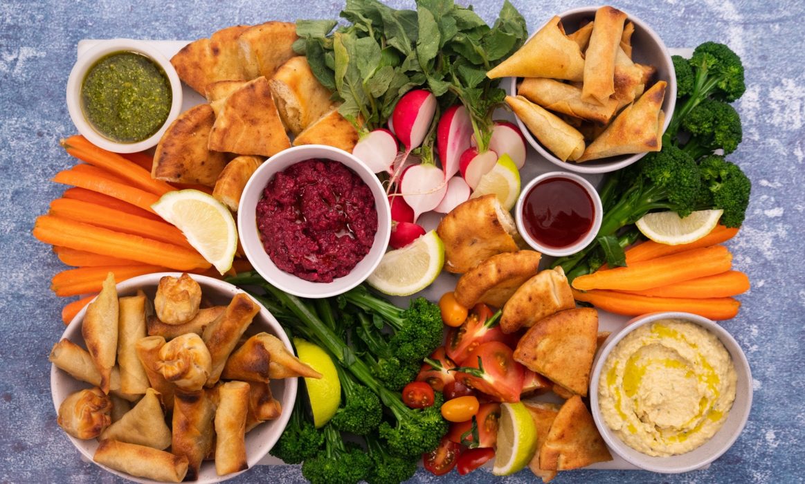 Small fried snacks, tomato sauce and hummus in small bowls on a rectangle tray among cut vegetables and lemon wedges.