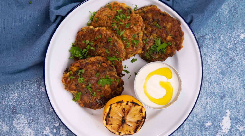 Four round fritters on a white plate with a pot of white sauce and charred lemon