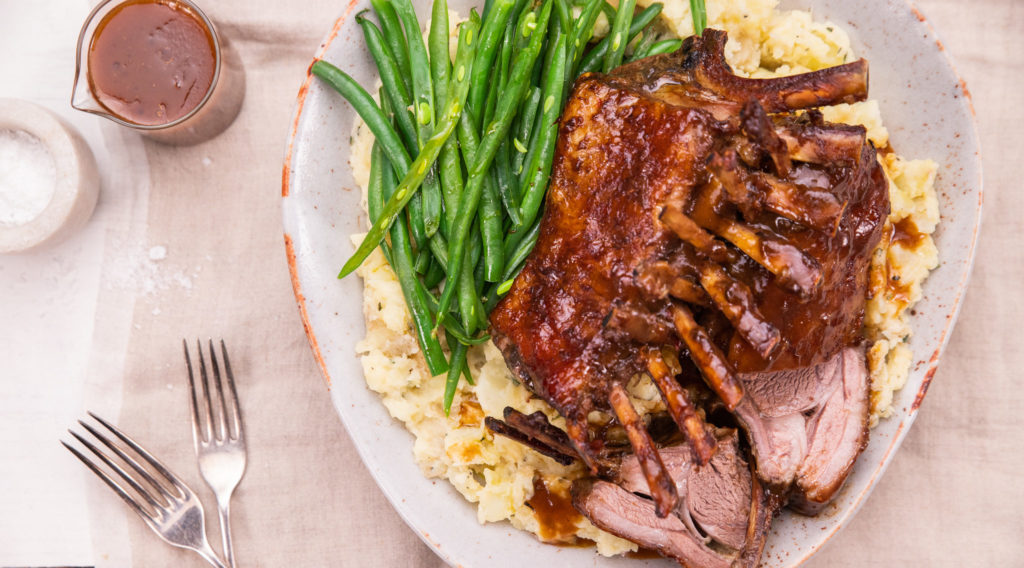 Cooked rack of lamb and green beans on large white bowl and two forks, two small pots of sauce and salt.