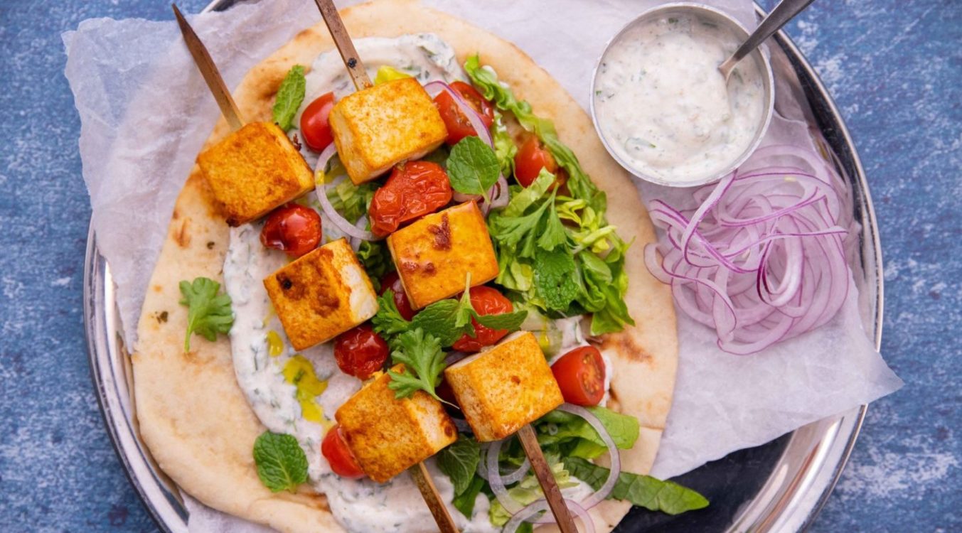 Two brown cube tofu and tomato skewers on bed of green salad on a flatbread with a small pot of white sauce