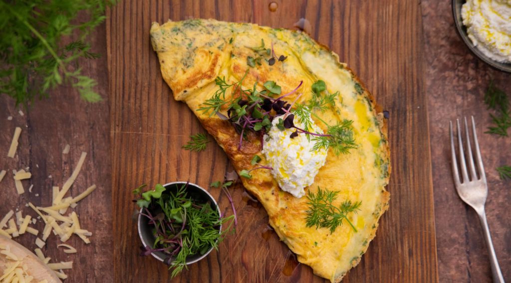 A half circle shaped yellow omelette topped with herbs and a blob of white cheese on a wood board and a small pot of greens and a fork