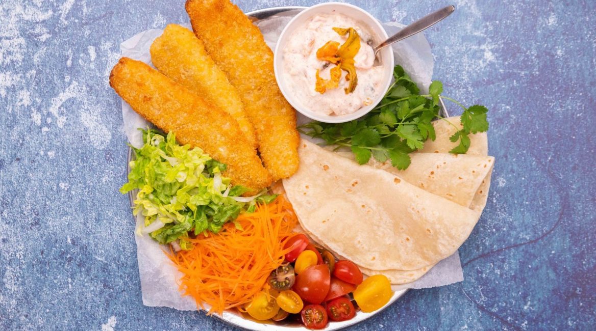 Crumbed fish, flatbread, tomatoes,greens and pot of white sauce on a plate on blue background