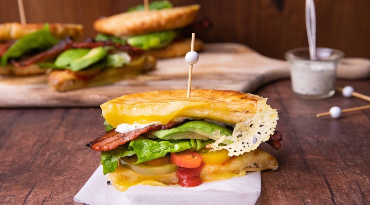 A side shot of toasted sandwich with avocado, bacon and tomato filling on a white board with some more in back ground.