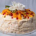 orange and red fruit pavlova with a white rose on top on a glass cake stand