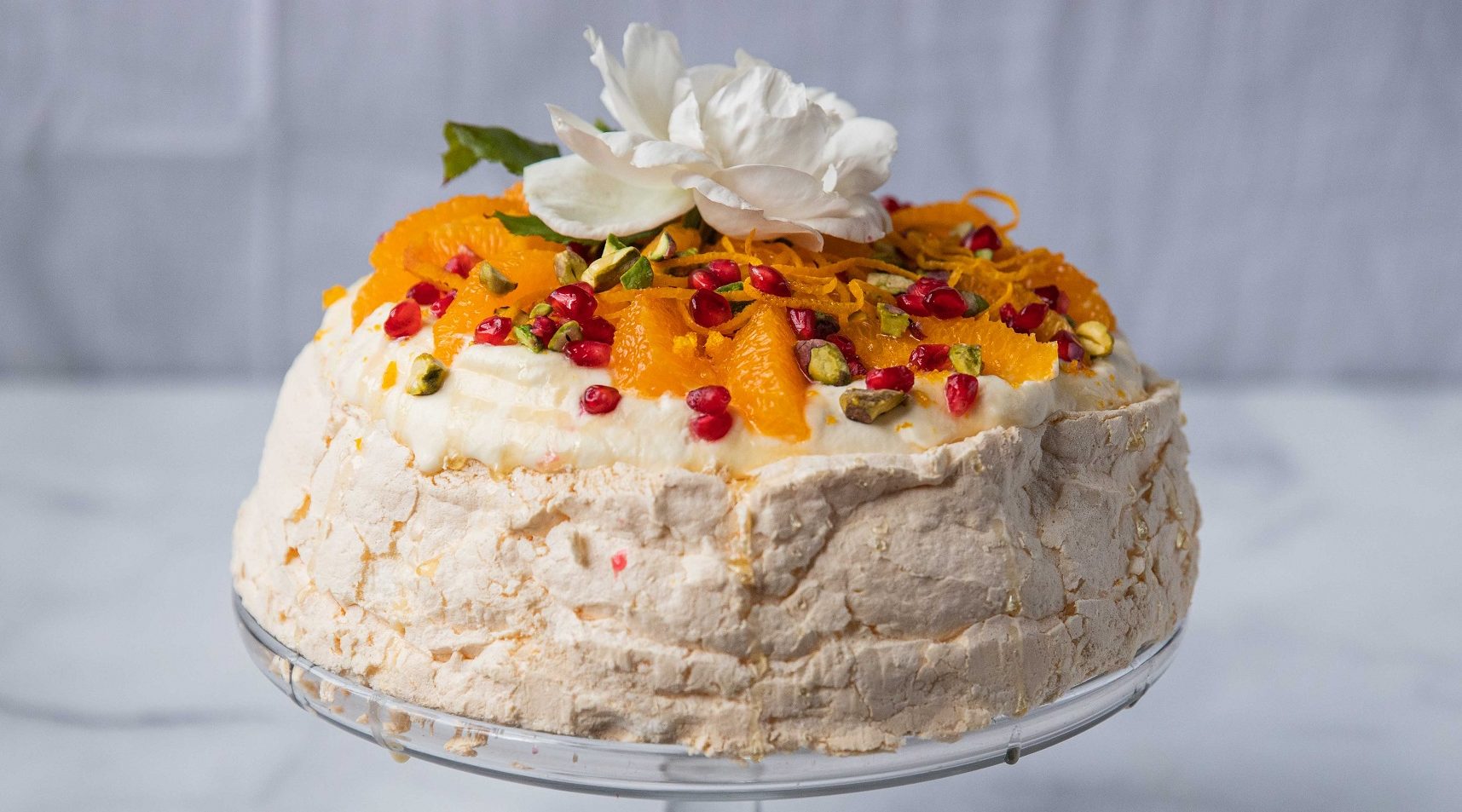 orange and red fruit pavlova with a white rose on top on a glass cake stand
