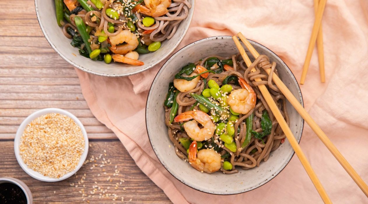 Two large bowls with noodles topped with prawns and greens with chopsticks and two small bowls on pink fabric.