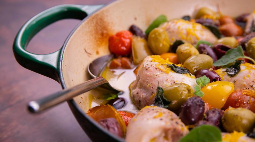 Chicken pieces, tomatoes and olives in a green pan with a handle on wooden board