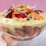 A glass bowl full of yellow cream and red and orange coloured fruit on pink & white background