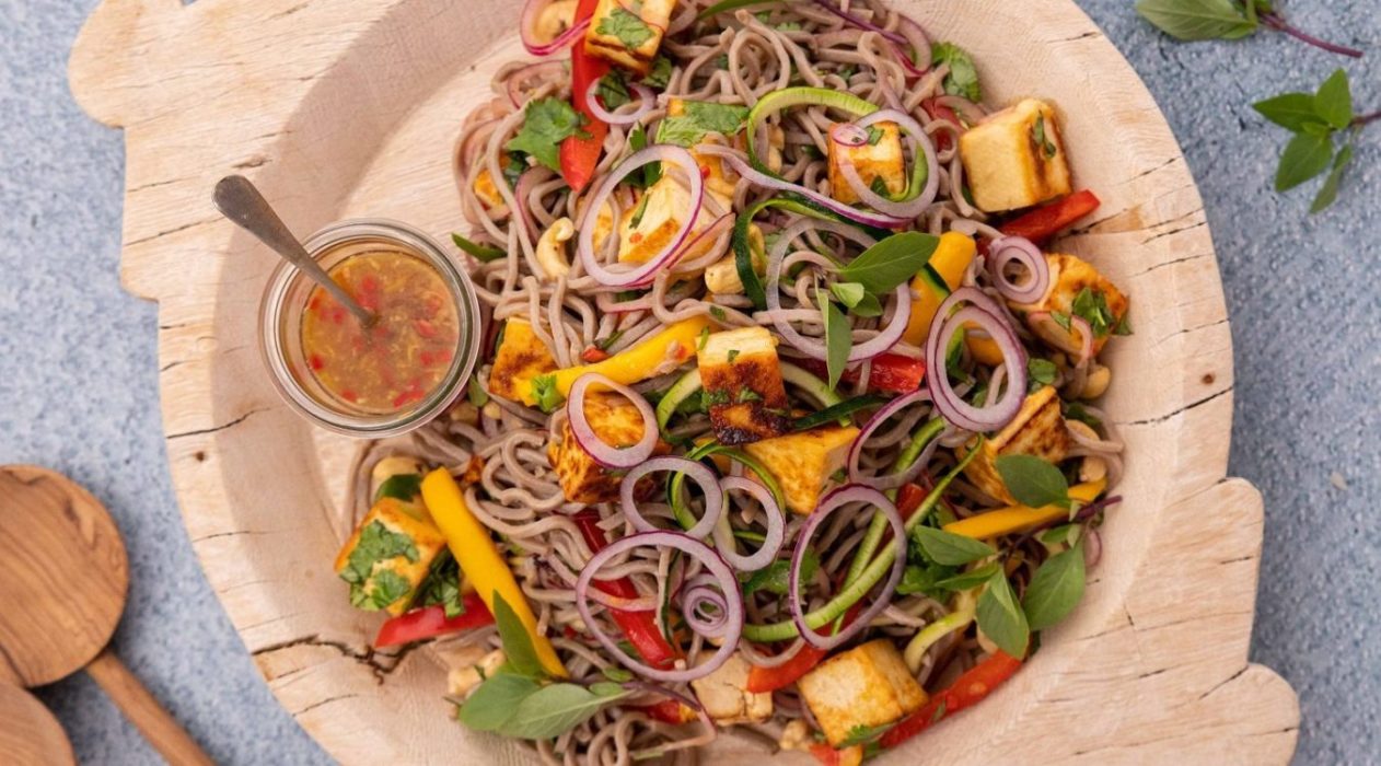 Gray noodles, white cubes, greens and red vege pieces on a wooden tray with serving spoons