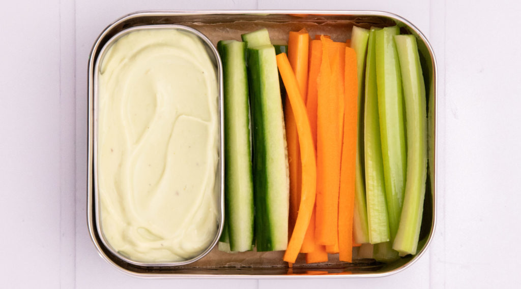 A pot of cream and cucumber,carrot,celery sticks in an oblong lunchbox on pale pink background.