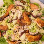Cooked chicken pieces and grated cheese on bed of lettuce on a wooden tray with a pair of salad server on blue background