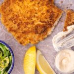 A piece of chicken schnitzel, a bowl of green salad, lemon wedges and a blob of mayonnaise and a knife on a wooden board