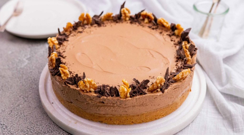 Coffee brown round cake decorated with walnuts and chocolate on white board on white fabric.