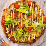 A colourful pizza on round marble surrounded by a chefs knife, mini tomato , shredded cheese and lettuce