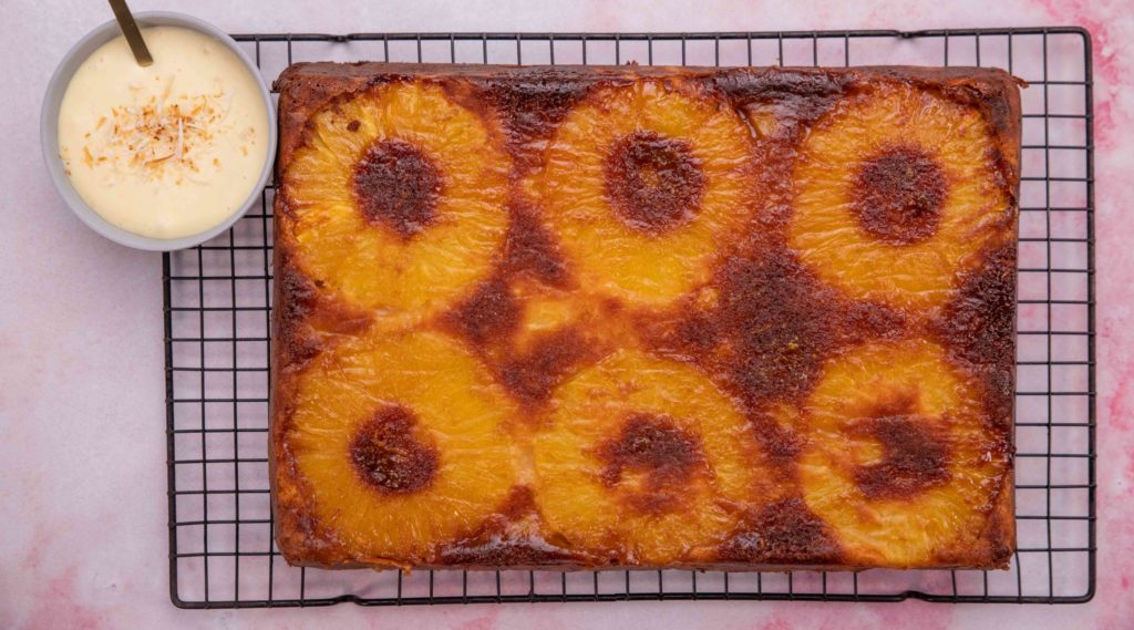 Large rectangle cake with 6 pinapple rings on wire rack with a small pot of cream
