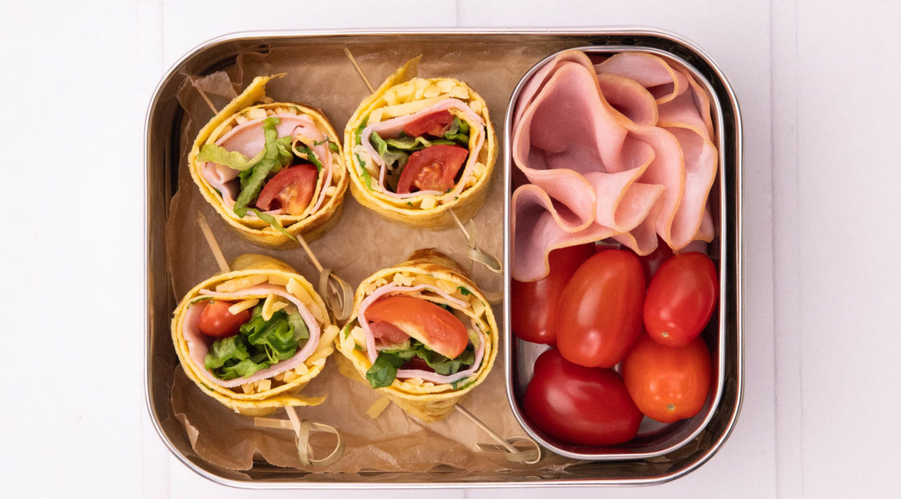 4 colourful rolled food with skewers, 4 mini tomatoes and some sliced ham in an oblong box on white board.