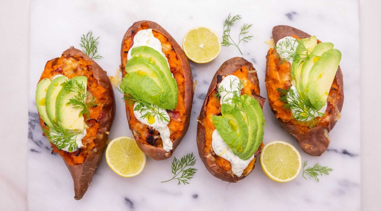 4 orange sweet potatoes topped with white cream and avocado slices, 3 lemon halves and bits of dill on white marble board.