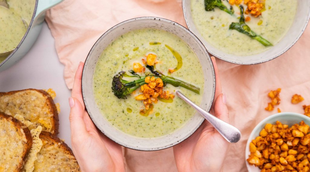 Hands holding a bowl of creamy green soup with brocoli and crumb on top with a spoon handle. A few cheesy toast and another dish of crumb with bits spilled on pink fabric.