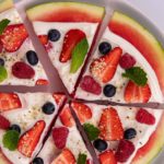 Round watermelon slice topped with white cream,berries and mint, cut into wedges on a round plate on white board