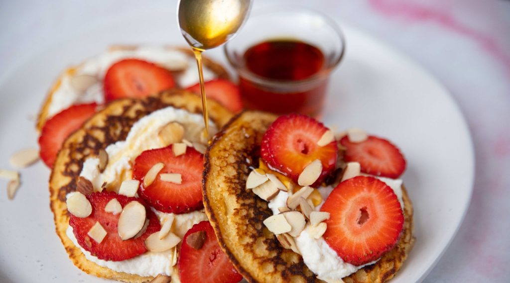 3 pancakes topped with white cream, strawberries and almond slices with a spoon pouring syrup and a small pot of it.