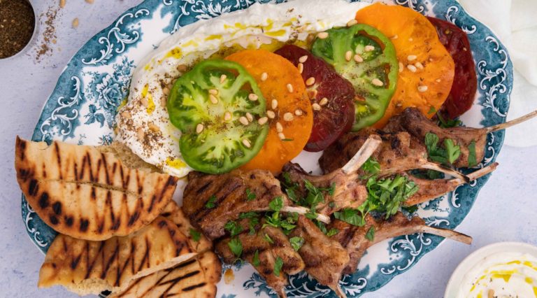 A platter with toasted pita bread, lamb cutlets, sliced green and red tomatoes on white sauce and nuts sprinkle.