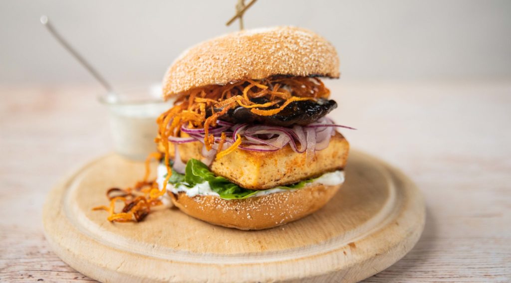 A burger with lettuce, square tofu, red onion, mushroom and fried carrot inside on a wooden round board and a small glass pot of white sauce at back. pot of